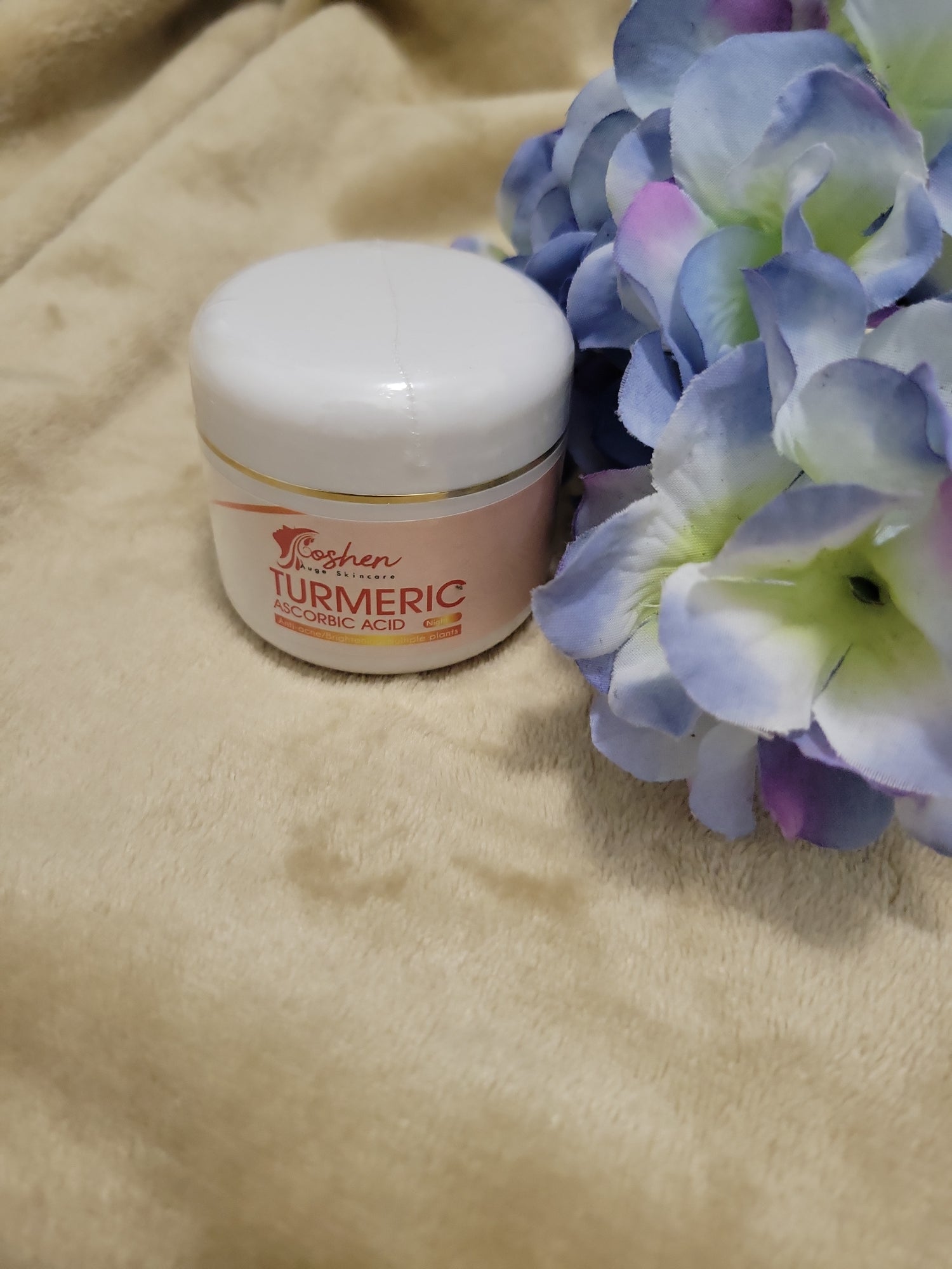 Turmeric Ascorbic Acid Cream jar next to blue and purple flowers on a soft surface.
